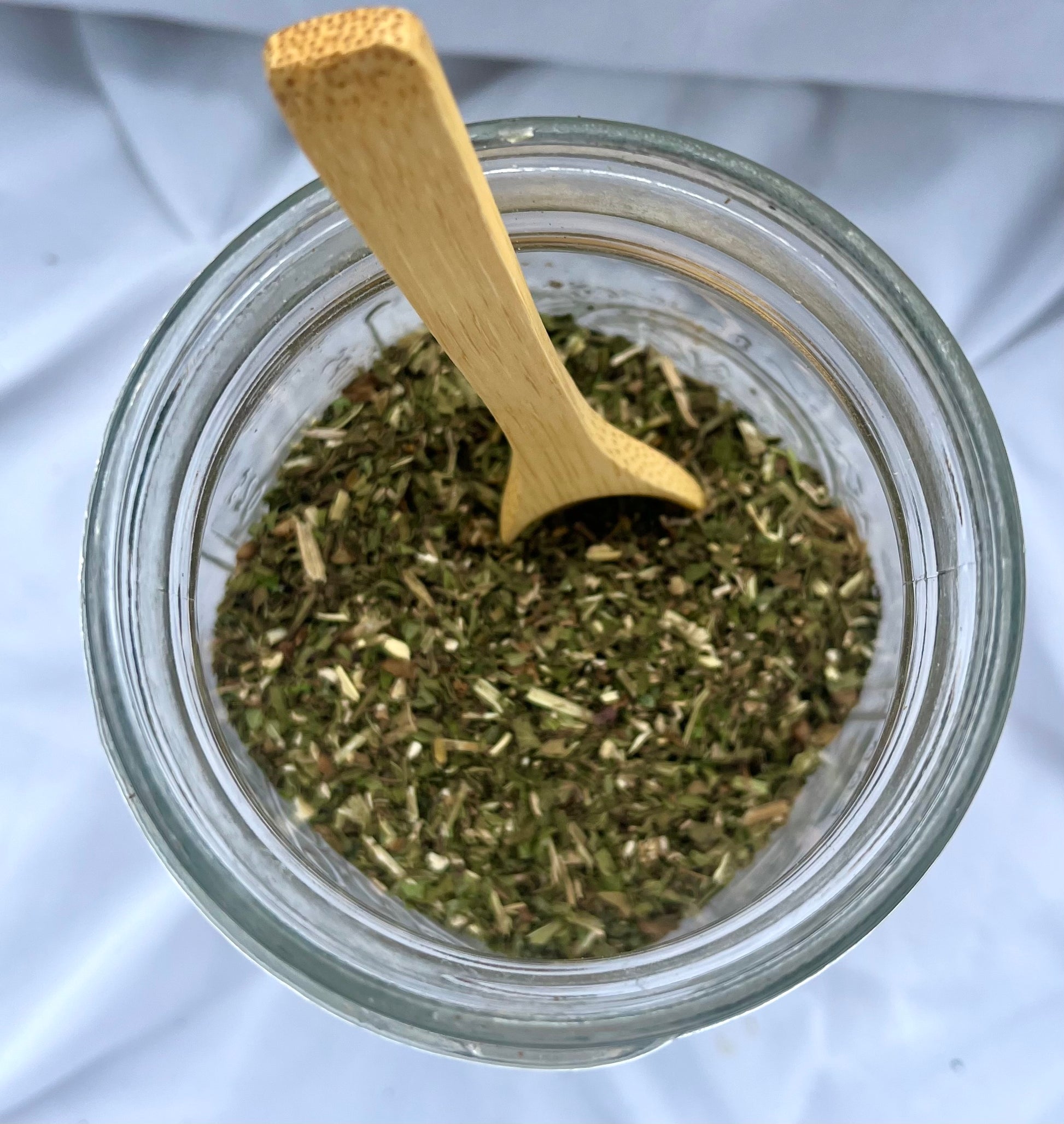 Glass jar filled with dried herbs and a wooden spoon on a white background