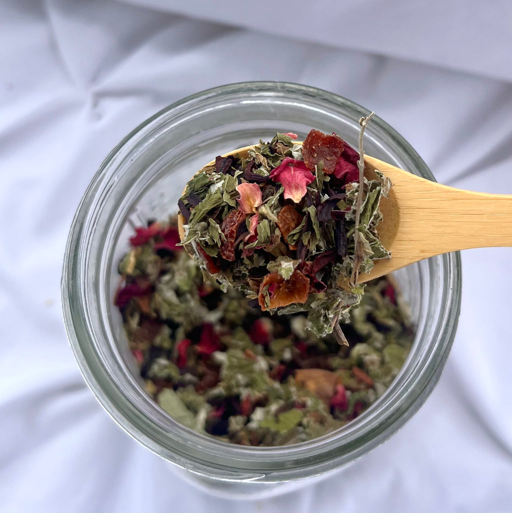 Glass jar filled with dried herbs and flowers, with a wooden spoon lifting some out.