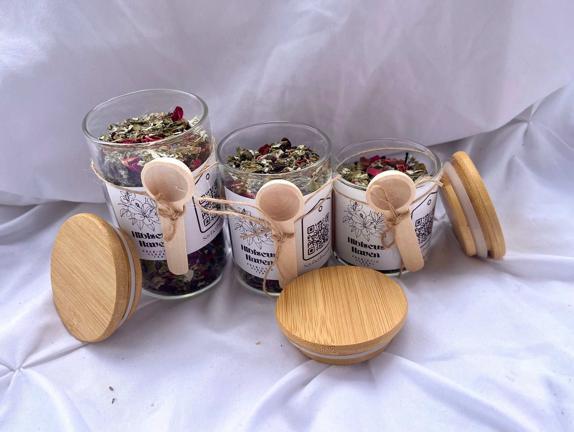 Three glass jars with wooden lids and spoons on a white fabric background