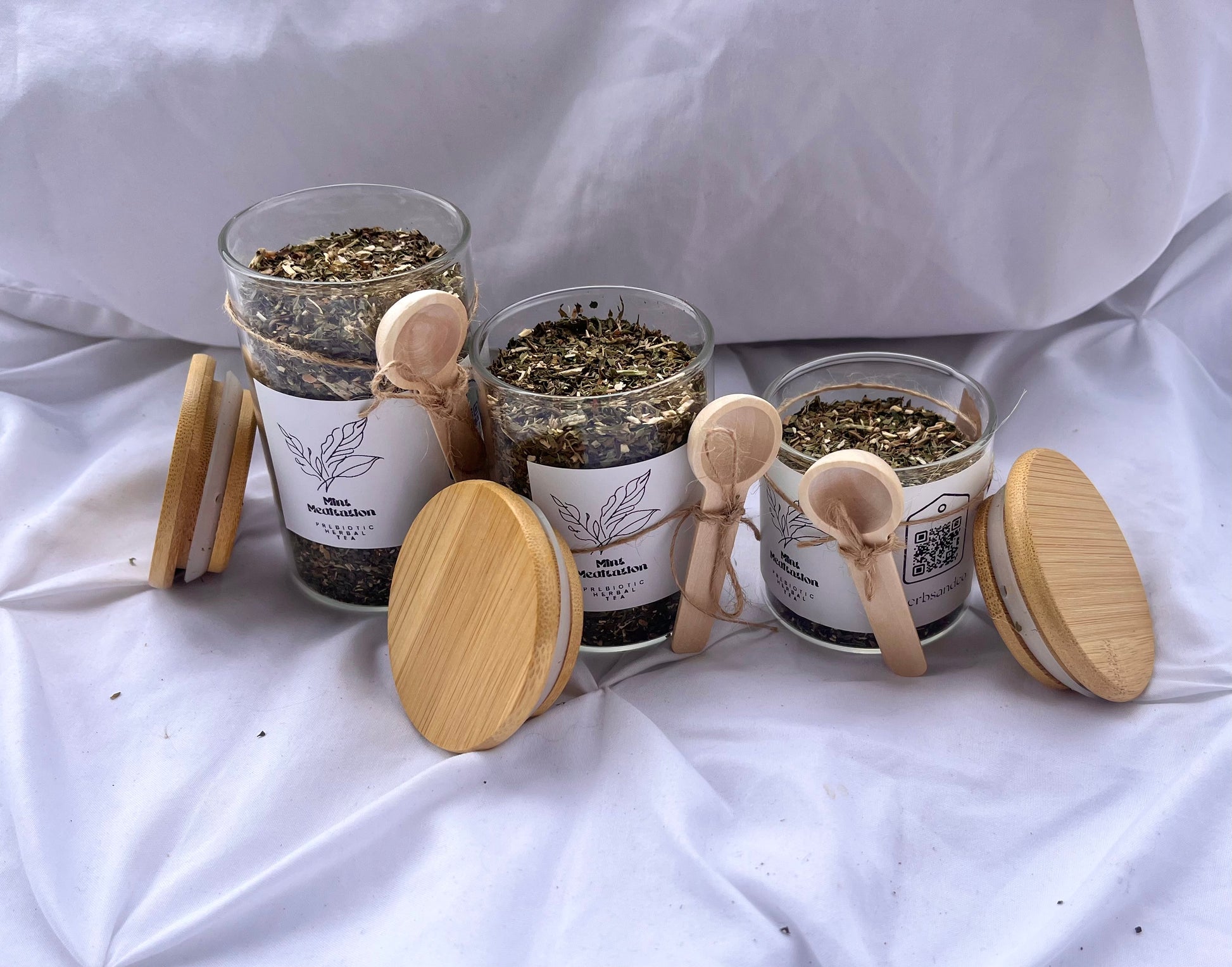 Four glass jars with wooden lids containing dried herbs on a white fabric background